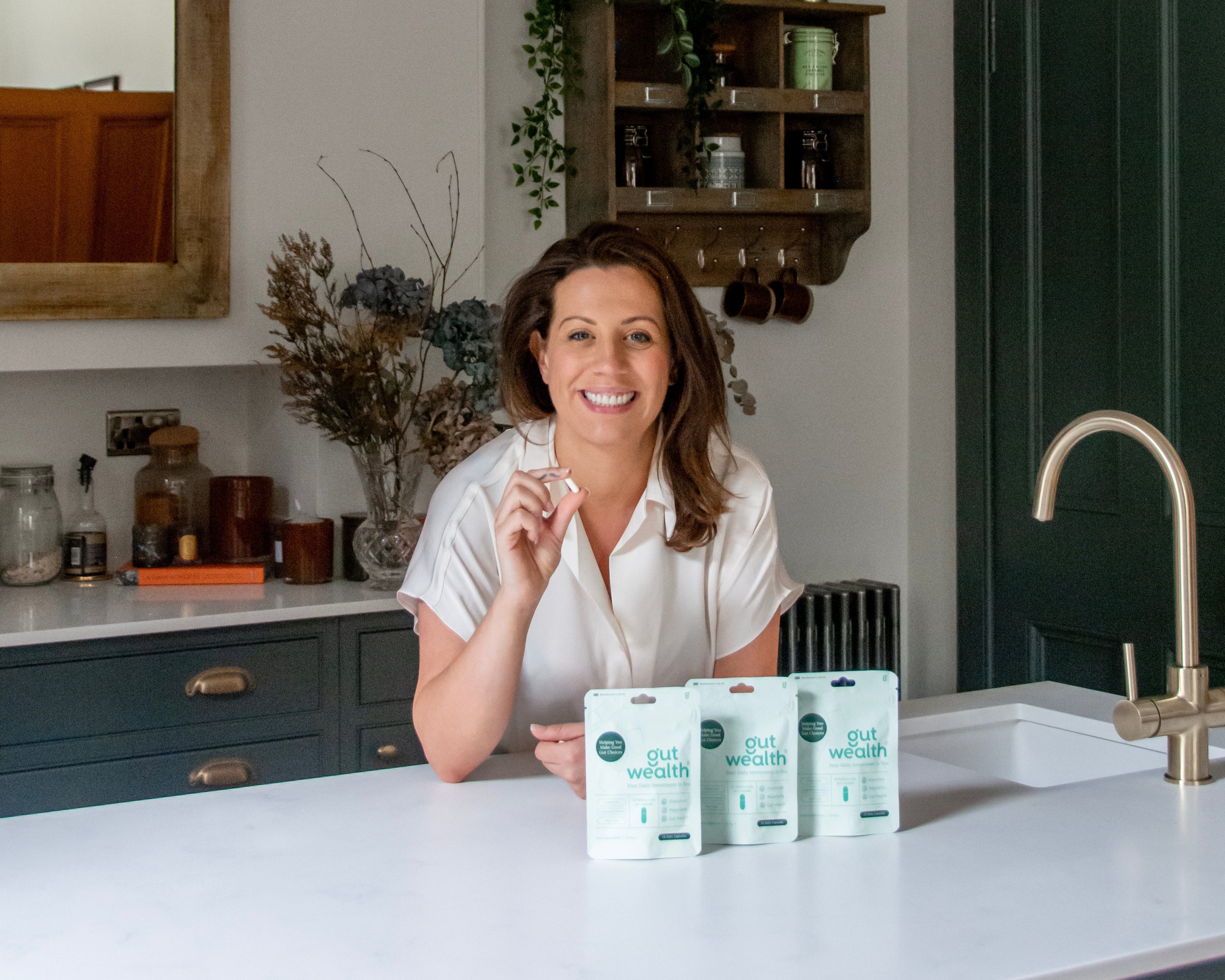 Woman in a kitchen holding a Gut Wealth Capsule, with 12-week supply of Gut Wealth Capsules on the counter.